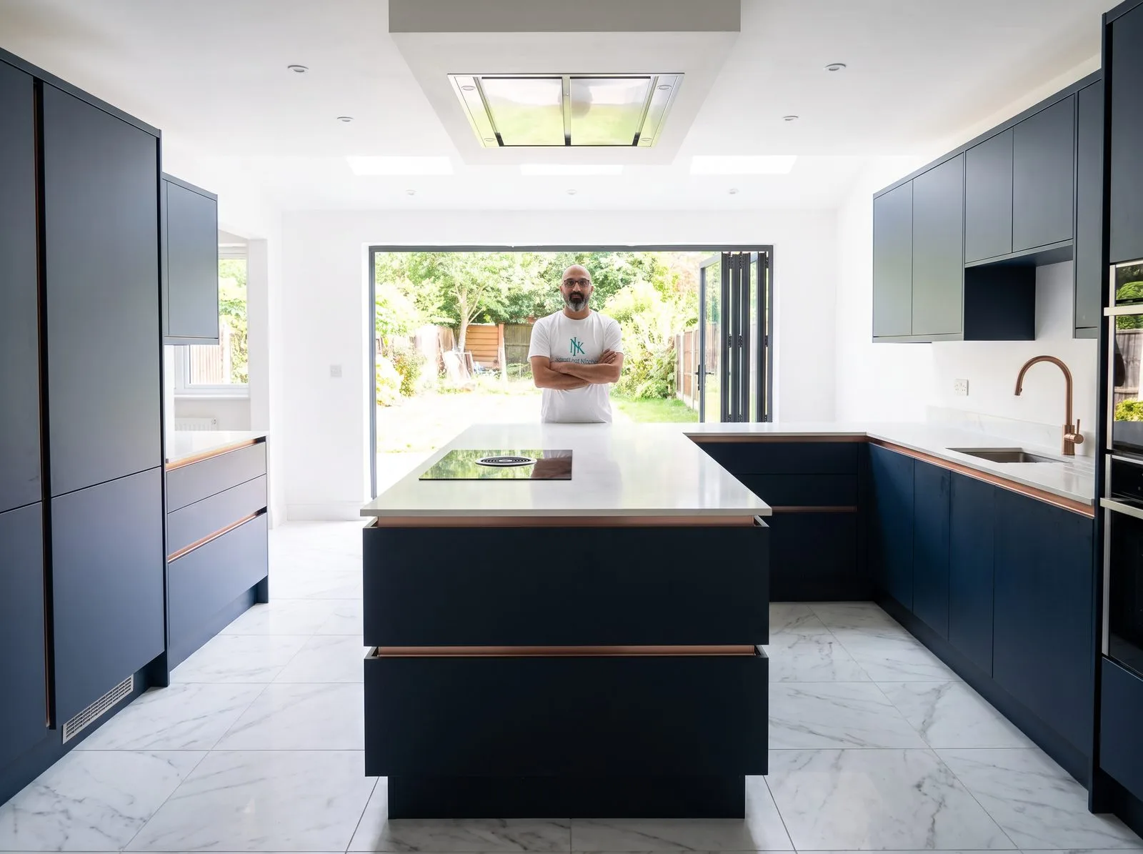 Finished U-shaped Wren kitchen with island, dark blue handleless cabinets and rose gold rail, bifold doors open